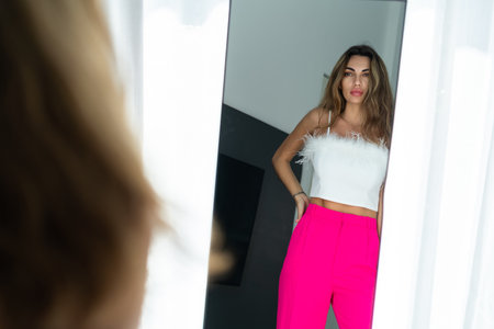 Stylish fit tanned beautiful woman in fashion pink pants and top posing in mirror at home bedroom, preparing for party, dressing upの写真素材