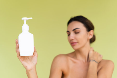 Beauty portrait of young topless woman with bare shoulders on green background with perfect skin and natural makeup holding bottle of shampoo, body lotionの写真素材