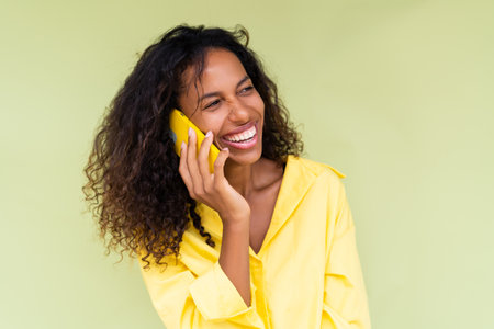 Beautiful african american woman in casual shirt on green background talk on mobile phone smile and laughの写真素材
