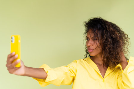 Beautiful african american woman in casual shirt on green background happy taking photo selfie on smart phoneの写真素材