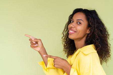 Beautiful african american woman in casual shirt on green background happy positive excited point finger to the leftの写真素材
