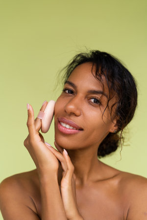 Beauty portrait of young topless african american woman with bare shoulders on green background with cosmetic powder puff velour makeup foundation blender sponge applicatorの写真素材
