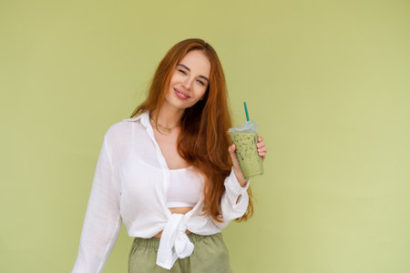 Beautiful red hair woman in casual shirt on green background positive vibes enjoying fresh ice green matcha tea with milk at hot summer dayの写真素材