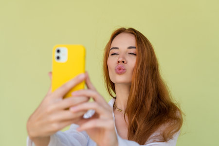 Beautiful red hair woman in casual shirt on green background happy taking photo selfie on smart phoneの写真素材