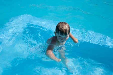 Emotional Toddler baby in the pool playing water toys. A little boy laughs by shooting water from a water gun in the camera.a little boy of 3 years old in an open-air pool with blue waterの写真素材