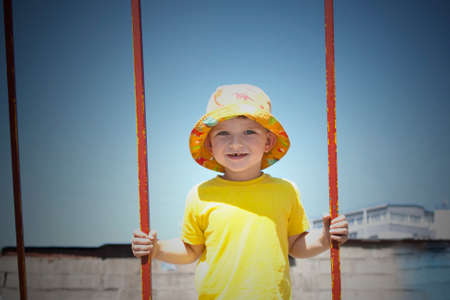 happy little boy 3 years old in a yellow T-shirt in a panama hat on a swing against the blue sky summerの写真素材
