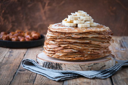 On the table on a stand, made of wood, cut a pile of thin pancakes, sprinkled with jam, sprinkled with powdered sugar, on top are banana and peanuts. In the background. Wooden background. Style Rusticの写真素材