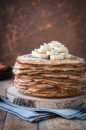On the table on a stand, made of wood, cut a pile of thin pancakes, sprinkled with jam, sprinkled with powdered sugar, on top are banana and peanuts. In the background. Wooden background. Style Rusticの写真素材