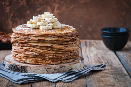 On the table on a stand, made of wood, cut a pile of thin pancakes, sprinkled with jam, sprinkled with powdered sugar, on top are banana and peanuts. In the background. Wooden background. Style Rusticの写真素材