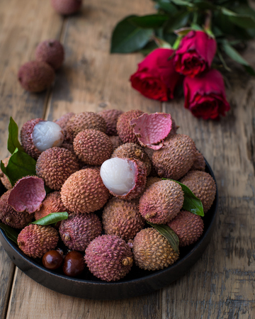 Still life of fresh lychee fruits in a black plate on a wooden background. Rustic style. Decorated with red rosesの写真素材