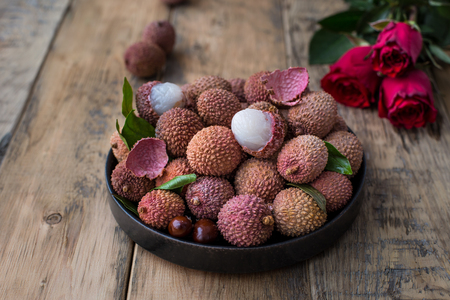 Still life of fresh lychee fruits in a black plate on a wooden background. Rustic style. Decorated with red rosesの写真素材