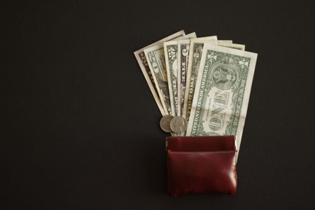 dollar banknotes and coins next to brown leather wallet on black background