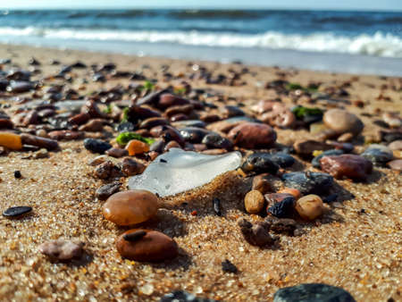 Macro shot of sea glass (beach glass) with smooth edges and accumulation of small pebbles in the sand of the beach of Baltic sea with visible water and waves in the backgroundの写真素材
