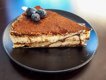 Triangular piece of italian dessert - tiramisu cake decorated with blueberries on a dark plate on...