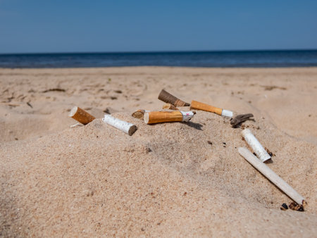 Different kinds of cigarette butts in the white sand on Baltic sea beach with water in background as toxic plastic pollution in the beach sand. Most littered plastic item in the world. Beach pollutionの写真素材