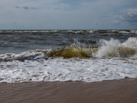 Close-up shot of big foamy waves in windy day of Baltic sea with brown sand on the coast and blue sky above. Seascape depicting Baltic seaの写真素材