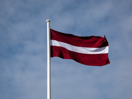 The national flag of Latvia in the wind with blue cloudy sky. Latvian flag - carmine red with white horizontal stripeの写真素材