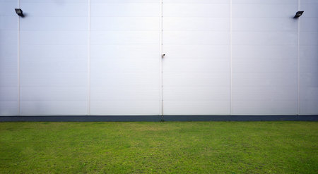 Wall of a warehouse building with lights and camera, grass field in foreground.