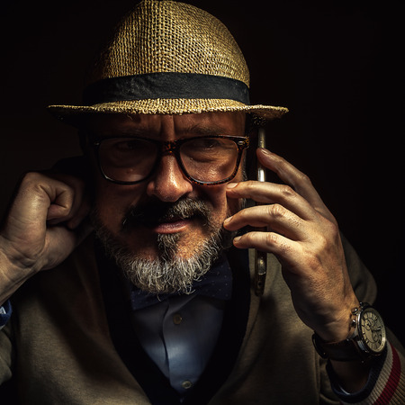 Older person communicating over cell phone, wearing retro style clothes, glasses and hat.の写真素材