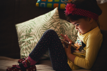 Little girl is sitting on couch and watching a mobile phone.の写真素材