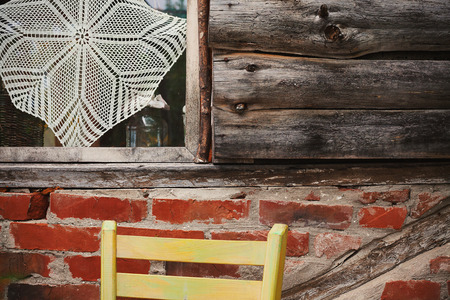 Closeup view of one part of an old wooden house, window with embroidery on one side, old weathered wooden planks on other and bricks in the bottom. Part of a wooden chair in front of a wall.の写真素材