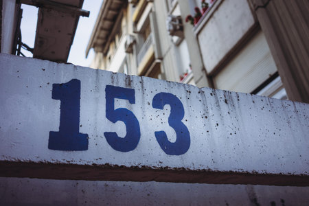 Number 153 painted with blue paint on a concrete wall, building in the background.