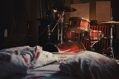 A red drum set next to an unmade bed, interior of the drummer's room by day.