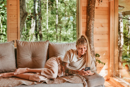 Woman with Smartphone Lying on Sofa in Log Cabinの写真素材