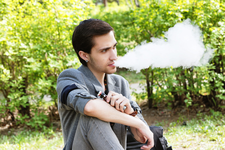 Thoughtful young businessman smokes a vape in a park in the spring afternoonの写真素材