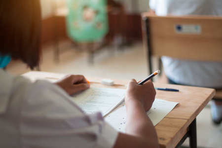 Students in uniform are doing the test in the classroom. Close up hand holding a pencil to write the...