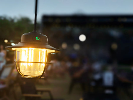 A glowing hanging lantern is in sharp focus at dusk, with warm light shining through the glass. The background shows blurred outdoor seating and people, creating a cozy atmosphere.の写真素材