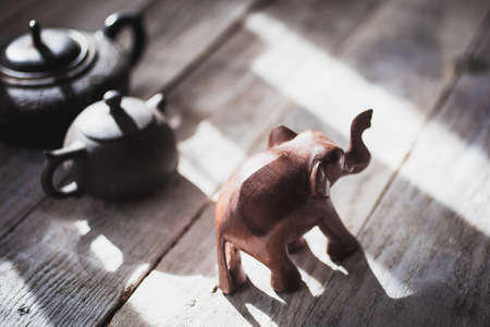 Decorative figurine of an elephant made of wood and teapots made of clay, on a wooden table. Bright sunlight from the window, hard shadowsの写真素材