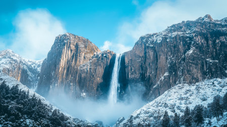 A stunning view of a waterfall pouring from rocky cliffs, surrounded by lush greenery and majestic snow-capped mountains, under a bright blue sky, perfect for nature lovers.の素材