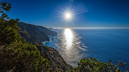 Explore this stunning coastal landscape showcasing the sun's radiant reflection on the tranquil ocean waters, bordered by majestic mountains under a clear blue sky.の素材