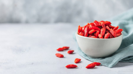 A beautiful bowl filled with vibrant red goji berries sits gracefully on a light grey surface, accompanied by a soft blue cloth, symbolizing health and wellness.の素材