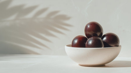 A serene composition of glossy dark fruits piled elegantly in a minimalist bowl against a light backdrop, enhanced by soft shadows, perfect for food art.の素材