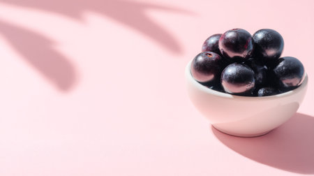 A visually appealing arrangement of fresh black grapes in a white bowl on a soft pink background, casting gentle shadows, perfect for healthy snack ideas or food presentations.の素材