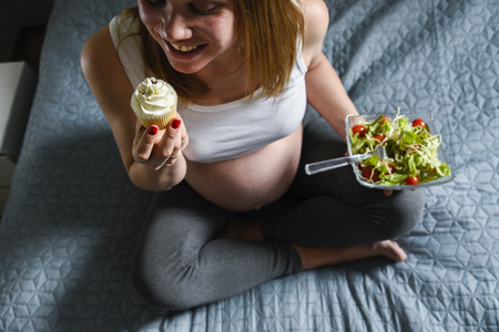 Pregnant woman holds green salad with tomatos in one hand and eating tasty sweet cupcakeの写真素材