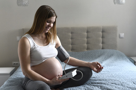 Pregnant woman cheking her blood pressure by herself sitting on bed in the room