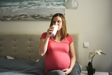 Young pregnant woman drinking milk sitting on bed in the roomの写真素材