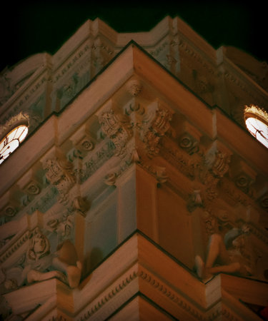 Dramatic night close-up of a historical building corner, showcasing ornate architectural reliefs and two brightly illuminated clock faces. Warm and cool tones. Ideal for concepts of time, history, urban mystery, or abstract architectural backgrounds.の写真素材