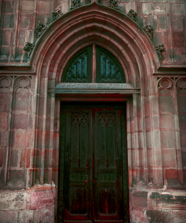 Imposing vertical shot of a dark, ornate wooden door set within a dramatic pink, red stone Gothic archway. Features intricate stone carving details and symmetry. Ideal for themes of history, religion, fantasy, secrecy, and European architecture.の写真素材