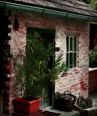 Close-up of a rustic, weathered red brick building exterior. Features a dark door, a window, and a potted plant next to a wicker basket. Captures cozy, nostalgic, and gardening atmosphere.の写真素材