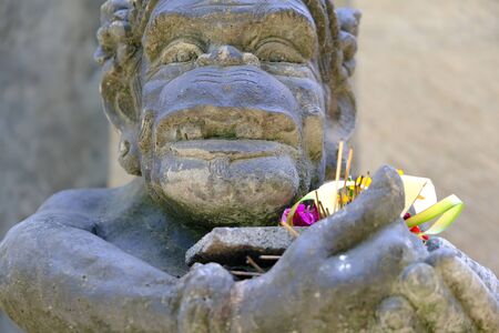 A statue of Balinese spirit holds offerings of palm leaves and flowers to the gods. Traditional Balinese sculpture.の写真素材