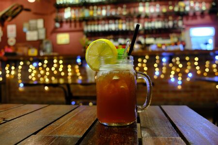 Iced tea in a mug decorated with a lemon slice and a black tube in the bar on a wooden table with a garland in the background. The concept of loneliness in a cafe.の写真素材