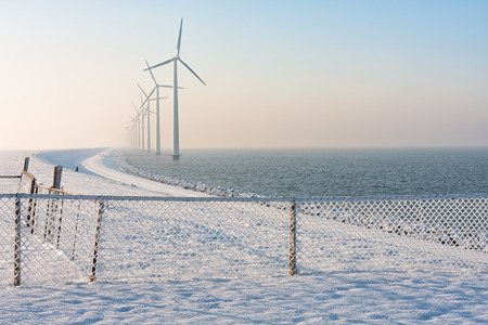 Dutch dike in winter with snow, fence and long row of wind turbines offshore in the water disapearing in winter hazeの写真素材