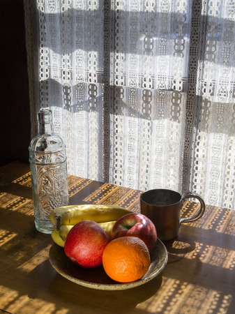 Bowl of fruit, empty bottle, and copper cup on a table in dappled light coming through a lace curtainの写真素材