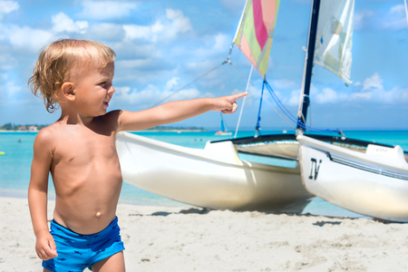 Cute blonde boy on the Carribean coast points his finger. Marvellous white yacht is on the background.の写真素材