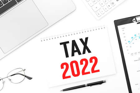 Text TAX 2022 on card. Laptop, glasses, pen, calculating machine and clipboard with charts and graphs. Business plan. Top view.の写真素材