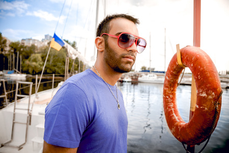 Closeup portrait of handsome latin man in sunglasses standing at seaport against yachts and lifebuoyの写真素材